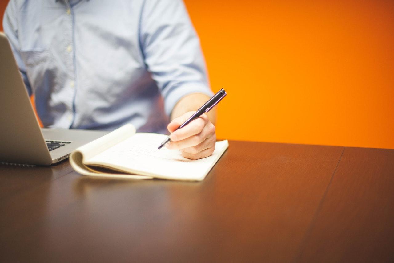 Person Writing on Note Beside Gray Laptop Computer